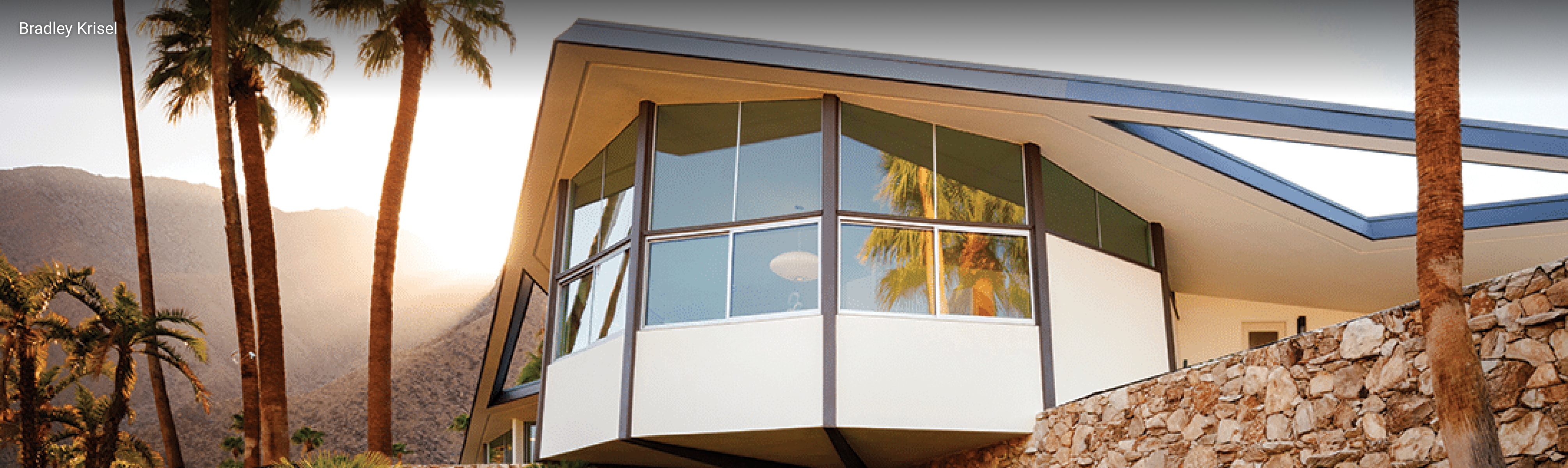 Mid-century modern home in Palm Springs, California featuring dramatic angular rooflines, cantilevered design, large glass windows, and stone exterior walls surrounded by palm trees and desert landscaping.