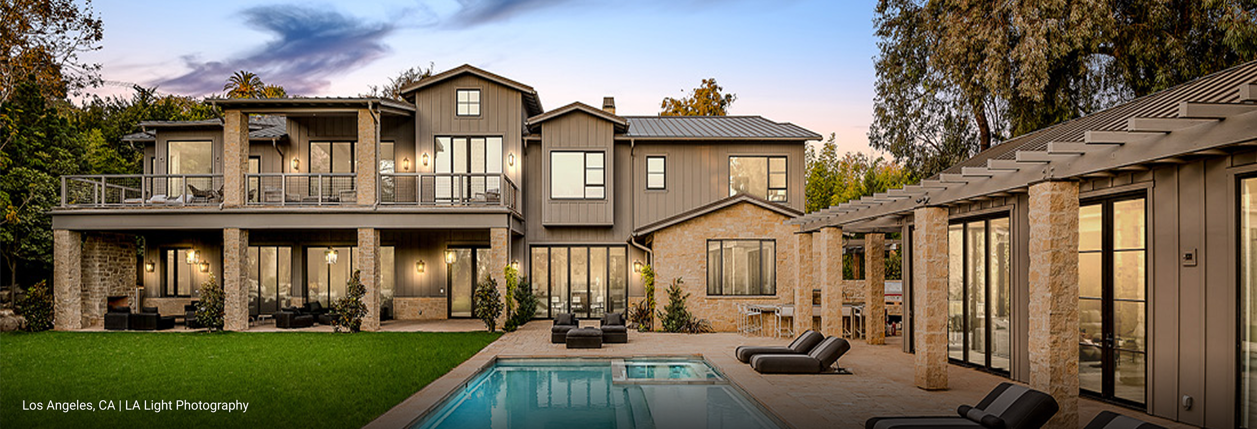 Luxury modern farmhouse in Los Angeles, California featuring multi-gabled roof design, gray board-and-batten siding, natural stone accents, and expansive glass doors opening to outdoor living space with pool and pergola.