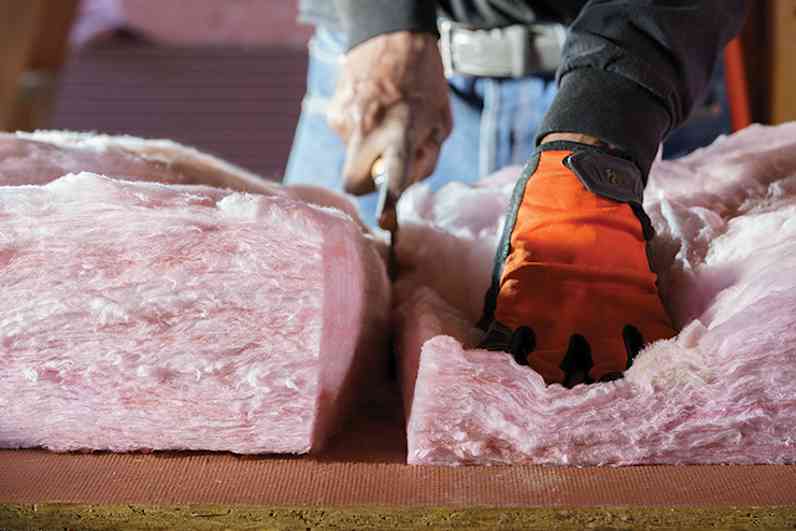 A professional insulation installer cuts PINK Next Gen™ Fiberglas™ with a large shear while wearing orange gloves.