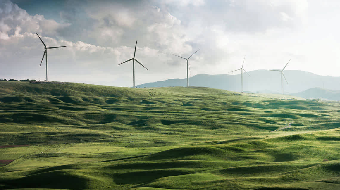 Wind Turbines in a hilly, grassy field with mountains int he distance.