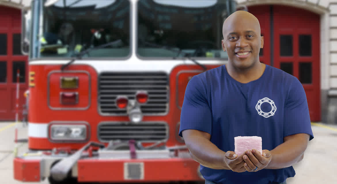 A smiling firefighter stands facing the camera in front of a fire station and a fire truck while holding a large cube of PINK Next Gen™ Fiberglas™ insulation.